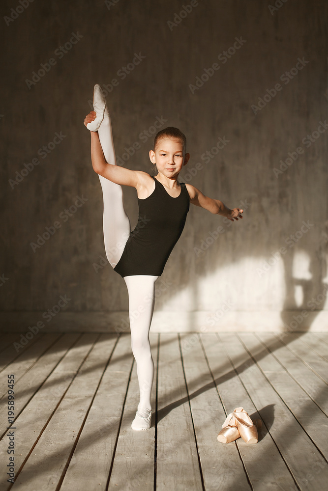 A little adorable young ballerina in black sportwear and white tights doing  vertical split in the interior studio posing on camera in bright sunlight  from window Stock Photo