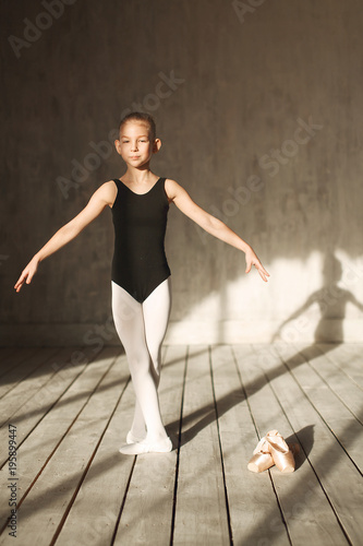 A little adorable young ballerina in black sportwear and white tights exercising in the interior studio posing on camera in bright sunlight from window