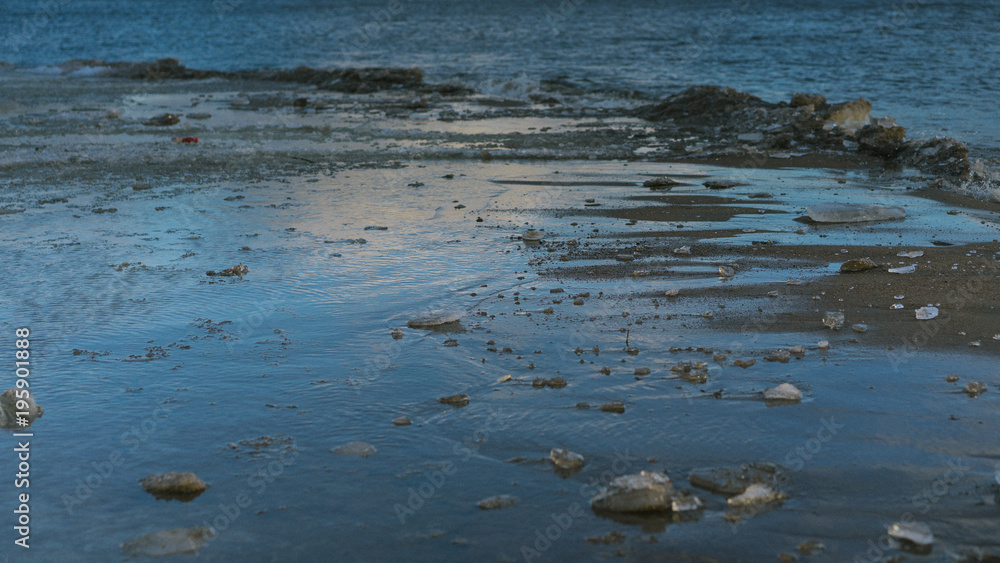 Close up picture of beach's surface. Ice is melted. Sand is wet and dirty. Some sun lights reflects on melted surface of ice cover.