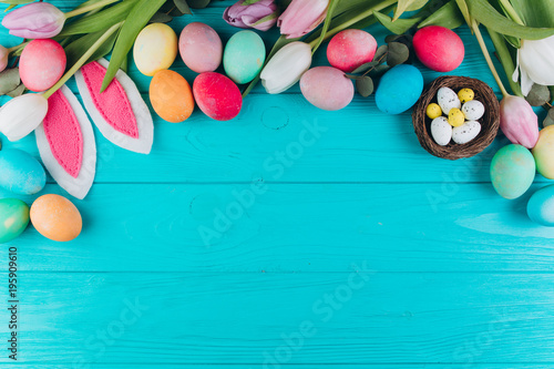 Easter composition with colored eggs, rabbit ears, nest and tulips on a blue wooden background.