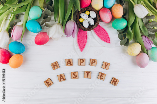 Wooden letters "Happy Easter" around colored eggs, rabbit ears, nest and tulips on a white background.