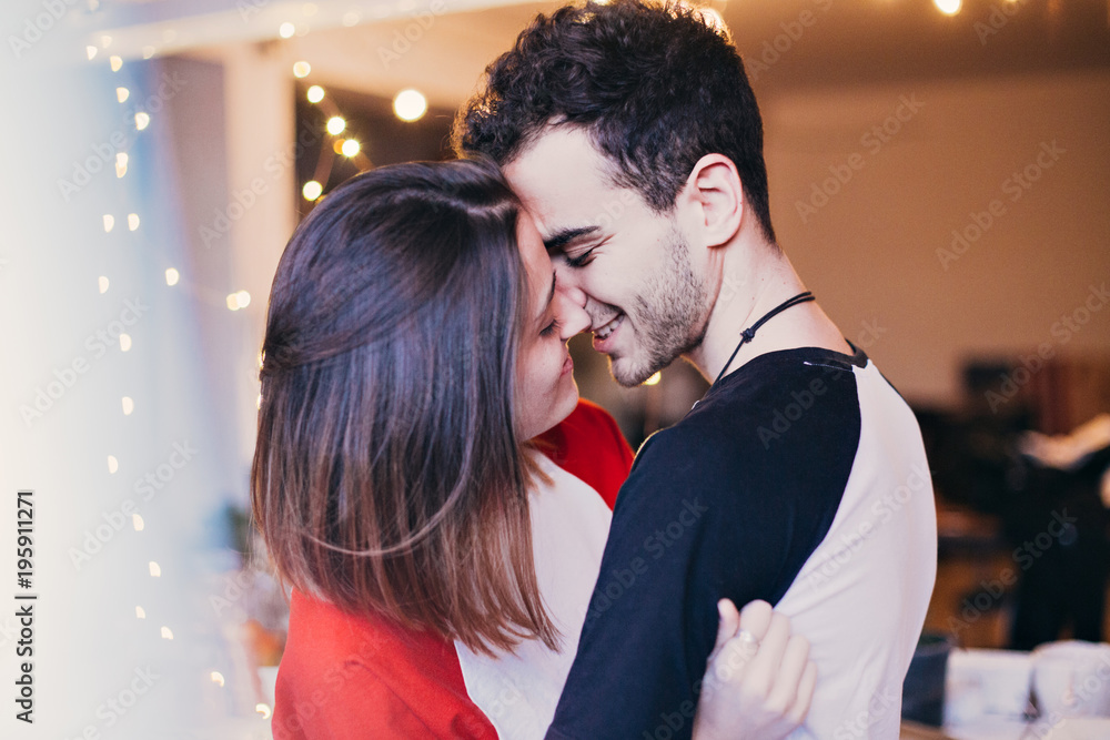 Cute and sensual young couple in love, smiling, hugging, cuddling and kissing inside house or apartment. Teenagers or millennials truly in love with each other, attractive boy and girl
