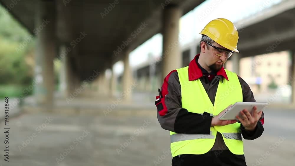 Workman with tablet computer under overpass. Manager of repair works at construction site under overhead road, inputs data in computer Workman with tablet computer under overpass. Manager of repair works at construction site under overhead road, inputs data in computer