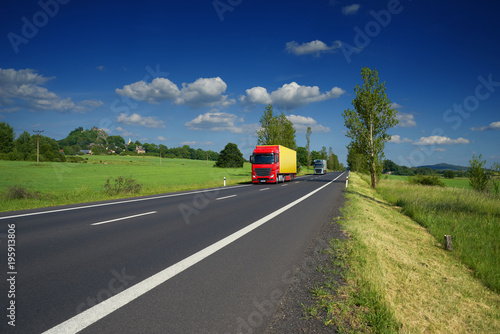 Trucks driving on the asphalt road in a rural landscape. Village and mountain with castle ruins in the background. photo