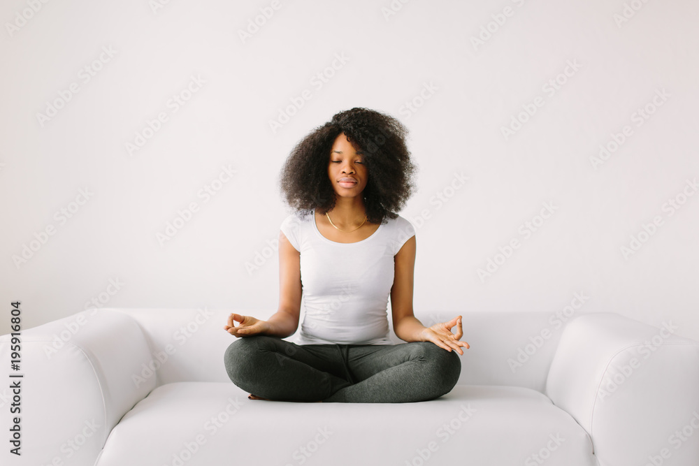 An African American young women sitting in the lotus position on white bed An African American young women sitting in the lotus position on white bed