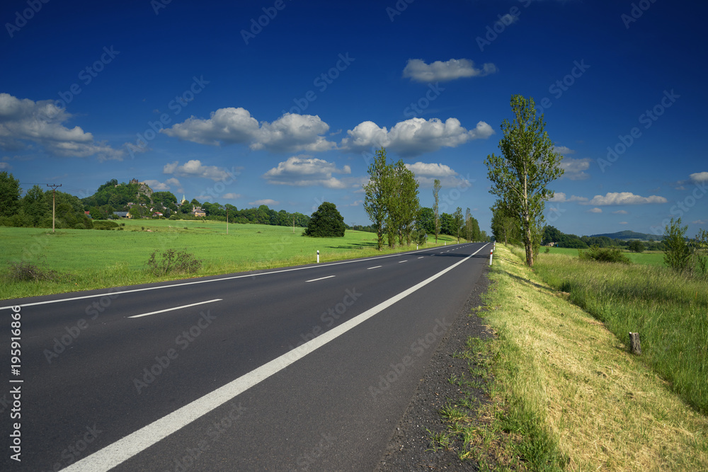 Empty asphalt road in a rural landscape. Village and mountain with castle ruins in the background. Empty asphalt road in a rural landscape. Village and mountain with castle ruins in the background.