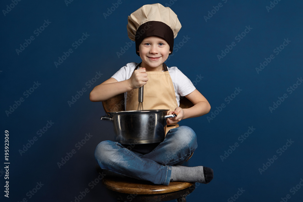 Isolated shot of happy cheerful male child in jeans, chef headwear and apron, sitting cross legged on wooden chair, holding casserole, beating up eggs with sugar while making dough for cake Isolated shot of happy cheerful male child in jeans, chef headwear and apron, sitting cross legged on wooden chair, holding casserole, beating up eggs with sugar while making dough for cake