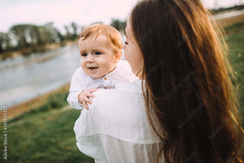 Cute little baby. Young beautiful mother is holding her baby in her arms walking with him on nature by the river Cute little baby. Young beautiful mother is holding her baby in her arms walking with him on nature by the river