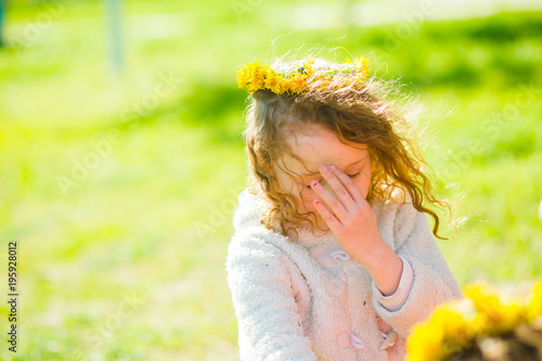 The girl in a wreath of dandelions covered her face with her hand. Child on a walk in the spring. The first rays of the sun. The first green grass.