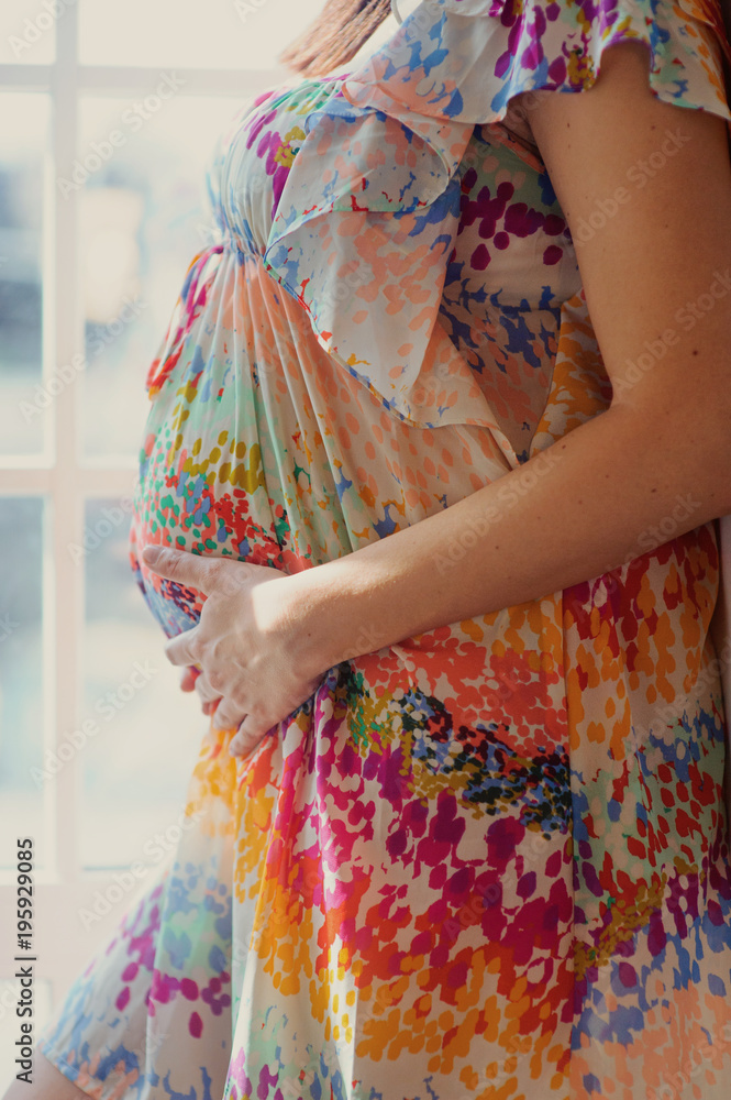 Pregnant woman in dress holds hands on belly on a white background. Pregnancy, maternity, preparation and expectation concept. Close-up, copy space, indoors. Beautiful tender mood photo of pregnancy.