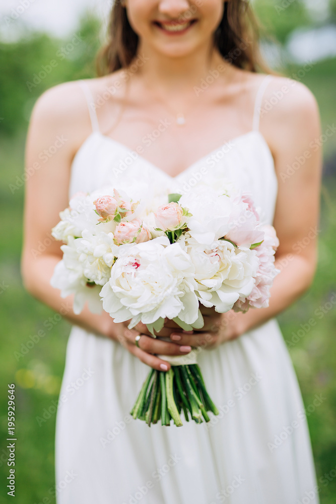 with a curly hand, the bride in a white dress, with curly flowing hair, holding in her hand a wedding bouquet of white peonies and pink roses against the background of a meadow with a curly hand, the bride in a white dress, with curly flowing hair, holding in her hand a wedding bouquet of white peonies and pink roses against the background of a meadow