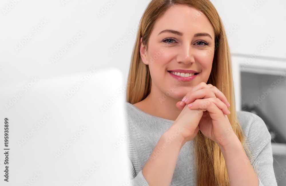 An attractive woman is looking at the camera while sitting at the desk in the workplace or at home. Sitting at the desk with a laptop. An attractive woman is looking at the camera while sitting at the desk in the workplace or at home. Sitting at the desk with a laptop.