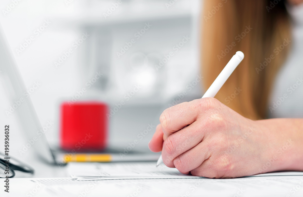 A woman writes while sitting at a table in an office or classroom. The girl fills the documents at the workplace using a laptop. A woman writes while sitting at a table in an office or classroom. The girl fills the documents at the workplace using a laptop.
