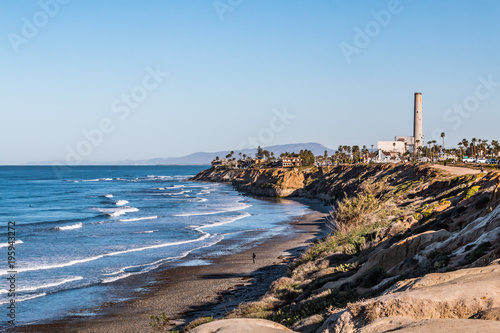 Man walks near cliffs on South Carlsbad State Beach in San Diego, California with the power plant landmark tower in the background. 