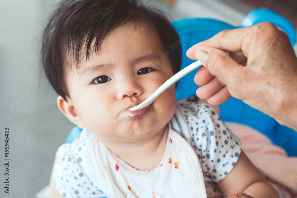 Grandmother feeding cute asian baby girl with a spoon at home. Little baby girl sitting in the chair and eating her meal with innocence. Grandmother feeding cute asian baby girl with a spoon at home. Little baby girl sitting in the chair and eating her meal with innocence.
