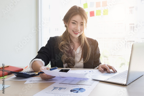 Young asian business woman using laptop computer and cell phone for contact customer on wooden desk in office. Business portrait concept.