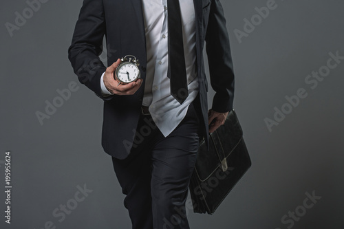 Close up of untidy tardy businessman. Shirt jerked from trousers. He carrying alarm clock and briefcase. Isolated on gray background