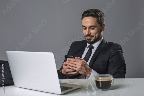 Smiling man sitting at table with laptop drinking coffee and surfing internet by phone