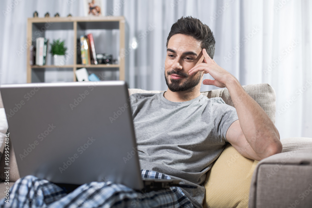 Portrait of pleased bearded man relaxing on sofa with notebook. Bookshelf on background Portrait of pleased bearded man relaxing on sofa with notebook. Bookshelf on background