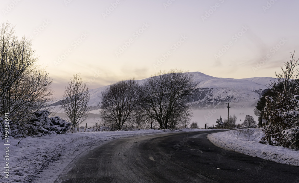 A road in the Scottish countryside in the Highlands surrounded by snow with mountains and a beautiful pink sunset in the background with clear sky ideal for copyspace A road in the Scottish countryside in the Highlands surrounded by snow with mountains and a beautiful pink sunset in the background with clear sky ideal for copyspace