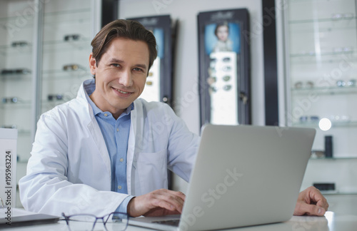 Portrait of beaming male ophthalmologist working with laptop while looking at camera. Optician store concept