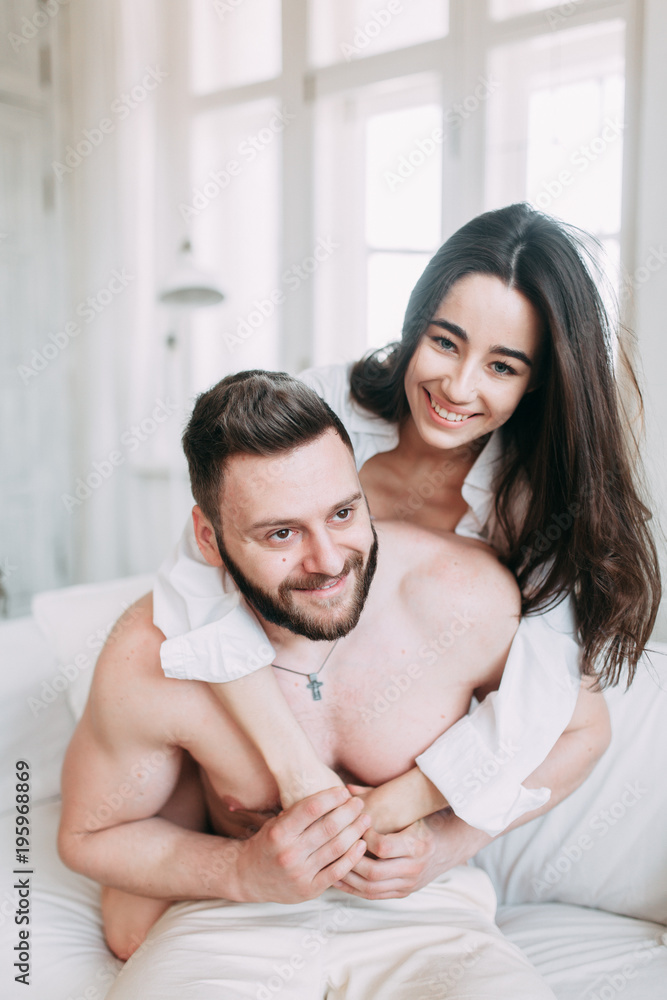 Young happy couple at home, cuddling, and kissing are. Beautiful Sunny morning with your loved ones in pajamas mad and laugh. Young happy couple at home, cuddling, and kissing are. Beautiful Sunny morning with your loved ones in pajamas mad and laugh.
