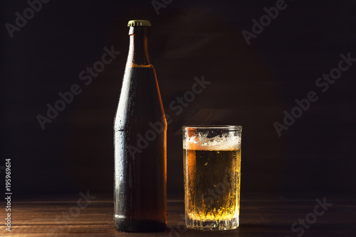 Cold beer bottle and glass with fresh beer in drops of water on dark background, craft brewery