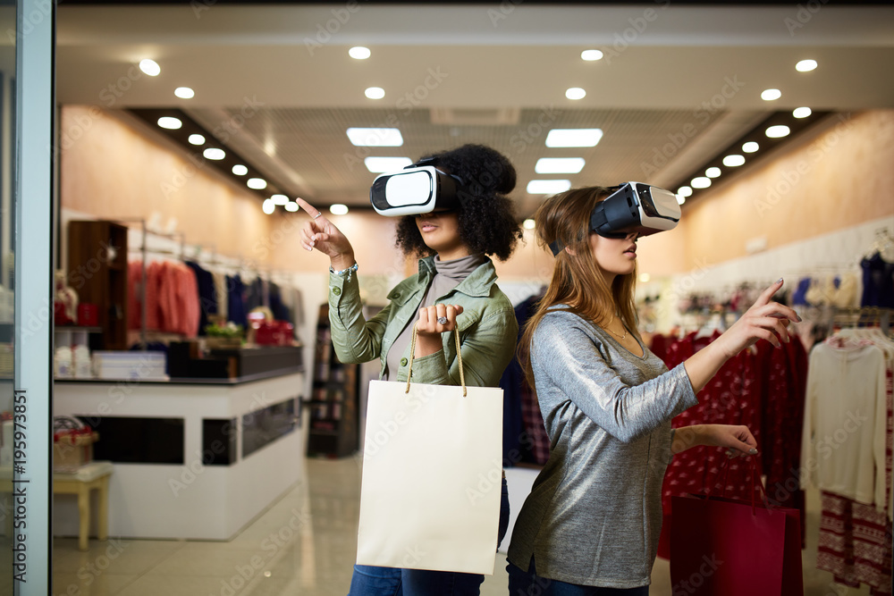 Two women in modern virtual reality headsets having expirience in shopping  at lingerie store. Multiracial girls in vr glasses with bags touching and  pointing interface elements in underwear shop. foto de Stock
