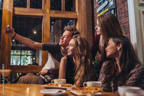 Excited friends taking selfie with smartphone sitting at table having night out.