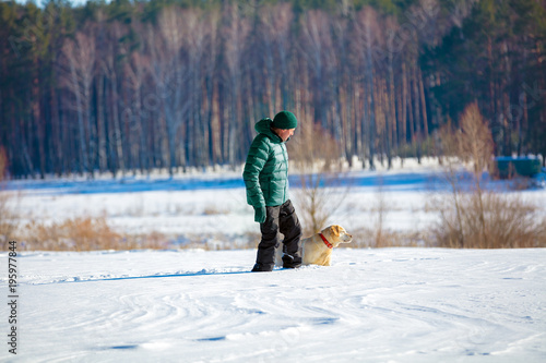 A man with a labrador retriever dog walking across a field covered with snow in winter