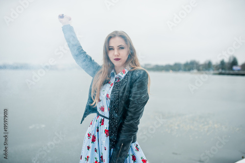 Stylish girl in leather jacket hold pink smoke flare at winter day against frozen lake.