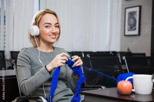 Young woman knitting at working place wearing headphones and smile. Leisure time at office concept. Businesswoman procrastinate