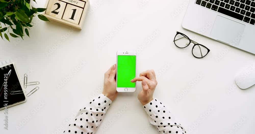 Top view on the white office desk and white smartphone with green screen and female hands taping on it. Vertical. Office stuff, laptop and glasses beside. Chroma key. Top view on the white office desk and white smartphone with green screen and female hands taping on it. Vertical. Office stuff, laptop and glasses beside. Chroma key.