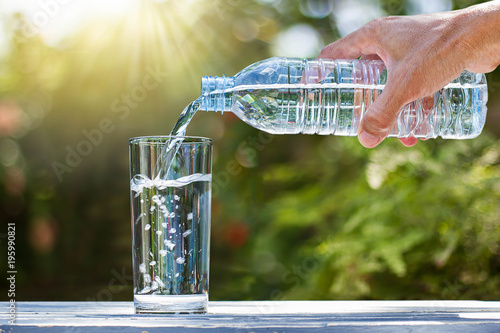 Hand holding drinking water bottle pouring water into glass on wooden table on blurred green nature background