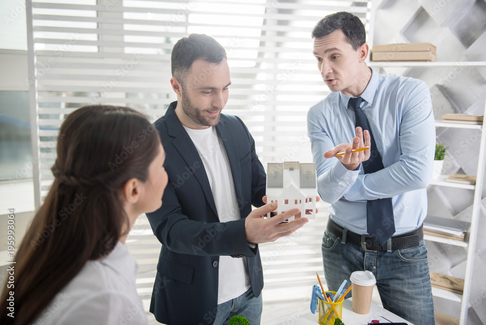 Future house. Handsome alert young dark-haired man smiling and holding a house miniature while his colleagues standing near him Future house. Handsome alert young dark-haired man smiling and holding a house miniature while his colleagues standing near him