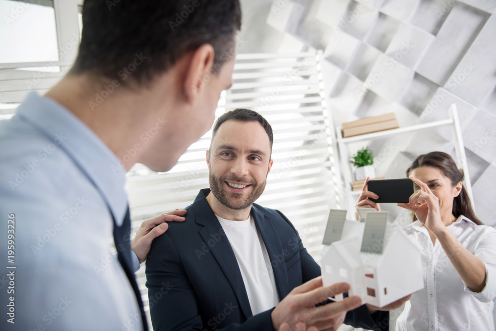 I feel happy. Attractive content young dark-haired architect smiling and holding a house miniature while their colleague taking photos of them I feel happy. Attractive content young dark-haired architect smiling and holding a house miniature while their colleague taking photos of them
