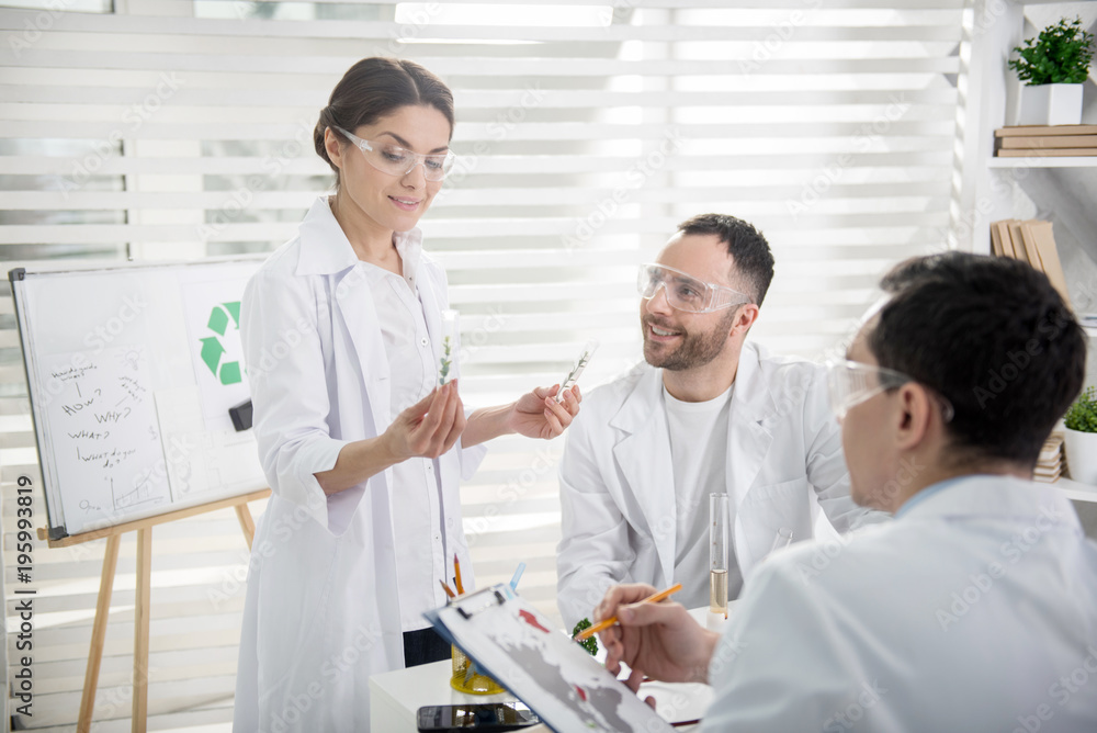 Best researchers. Beautiful content dark-haired ecologist smiling and holding test tubes and her colleagues sitting at the table Best researchers. Beautiful content dark-haired ecologist smiling and holding test tubes and her colleagues sitting at the table