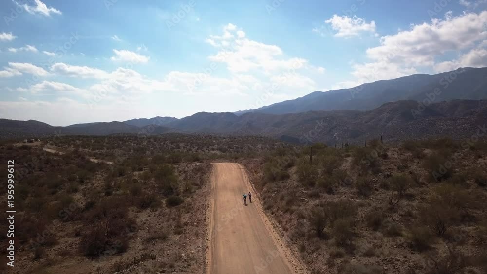 Two cyclists riding bicycles on a long dusty dirt road in a desert landscape. Two cyclists riding bicycles on a long dusty dirt road in a desert landscape.