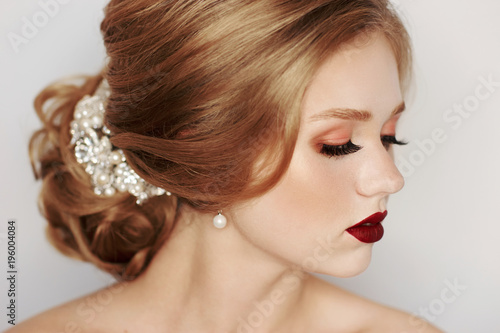 Beautiful young woman with blonde hair decorated with luxurious wedding accessory, pearl earrings and evening makeup. Portrait of attractive female model or bride with elegant hairstyle and red lips.