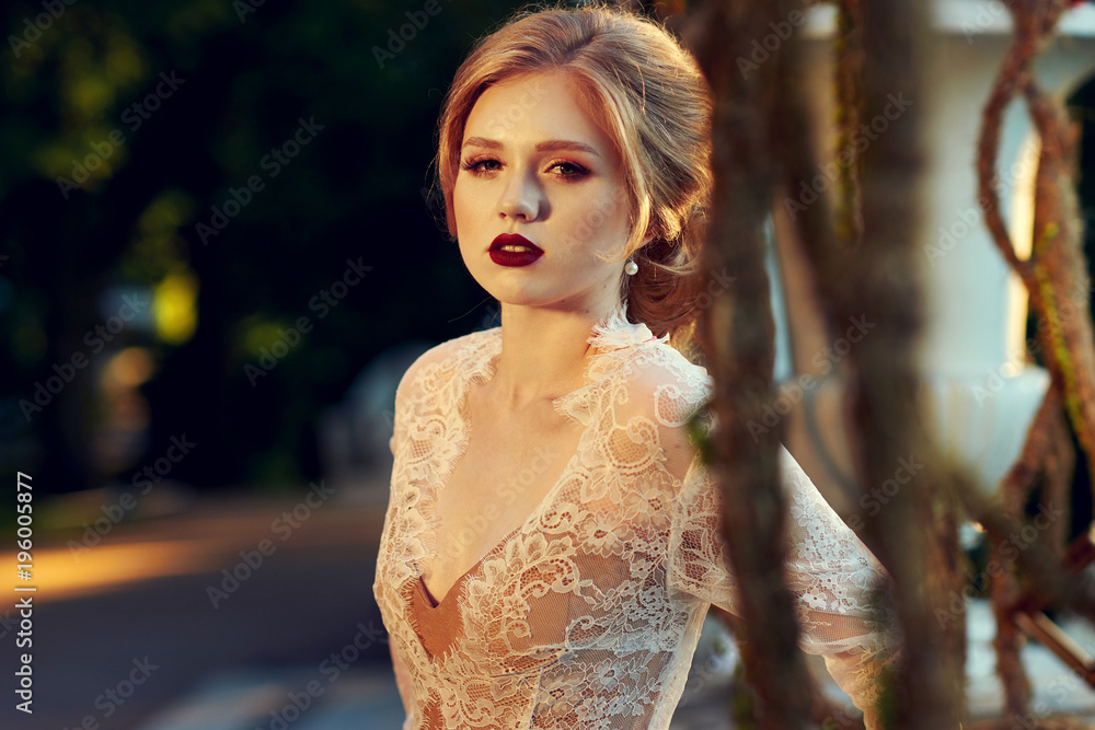 Naklejka premium Blonde woman with elegant hairstyle wearing dress and white lace jacket, standing on garden terrace and leaning on balustrade decorated with large flower vases against shining sun on background.