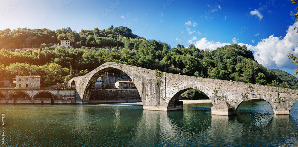 Fototapeta premium Picturesque view of medieval stone arch bridge Ponte della Maddalena across river Serchio in Borgo a Mozzano, Lucca, Tuscany, Italy. Scenic travel destination postcard.