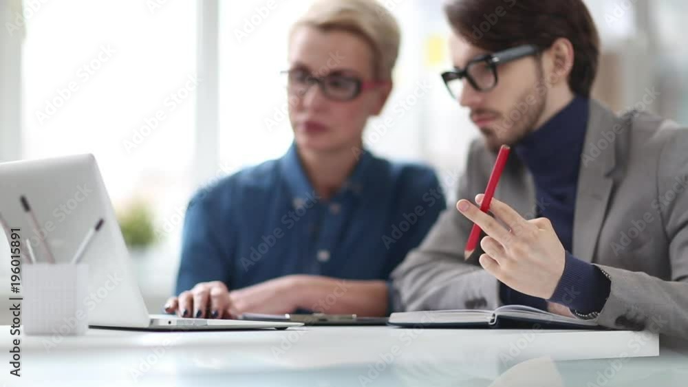 Joint work of talented young designers: pretty young woman sitting at office desk and working on ambitious project, her male colleague looking at laptop screen and twisting pencil Joint work of talented young designers: pretty young woman sitting at office desk and working on ambitious project, her male colleague looking at laptop screen and twisting pencil