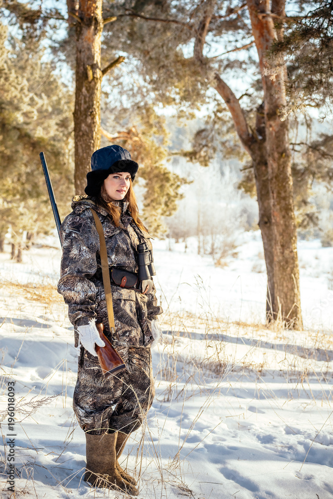 Portrait of a beautiful young girl while hunting in a winter pine forest, with a gun