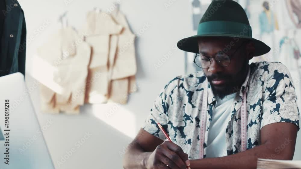 Handsome Afro-American designer in a cool outfit actively sketches his new fashion collection. Comfortable, light office. Skillfully, work of art, workmanship. Slow motion, male portrait Handsome Afro-American designer in a cool outfit actively sketches his new fashion collection. Comfortable, light office. Skillfully, work of art, workmanship. Slow motion, male portrait