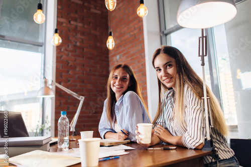 Portrait of two pretty smiling young women looking at camera sitting at work desk. Female freelancers working at home