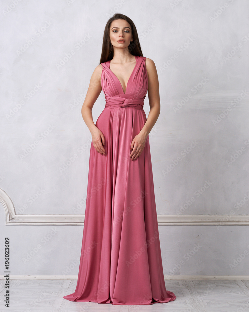 Beautiful long haired young woman dressed in stylish red bandeau maxi dress posing against white wall on background. Elegant brunette female model demonstrating evening outfit in studio.