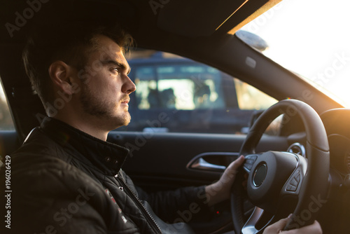 Concentrated young man driving a luxury car. Serious guy looking forward and holds hands on the wheel. Successful man in a hurry on business. Sun rays falling on face. Side view © Sergey_T