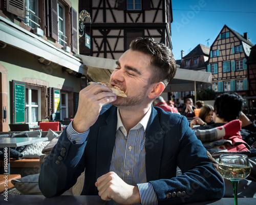 Very hungry man eats pizza in a cafe outdoors. Young guy in a suit is eating meal with wine. Terrace with beautiful view on vintage buildings in small European city