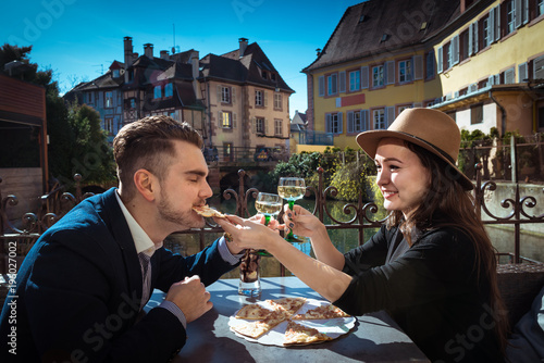 Young woman is feeding her boyfriend. Two stylish people sits together in local cafe with food and wine at city promenade. Trendy couple has lunch in Alsace. Man in the suit eats pizza with girlfriend