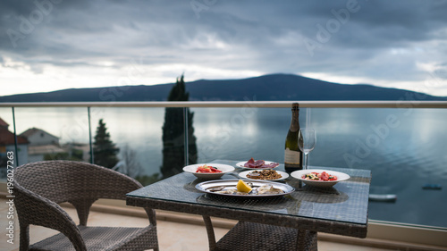 Table with local food and wine on the home balcony. Focus on food. Beautiful view from the balcony on calm sea and mountains. Oysters with lemon, salad, meat, snacks and bottle of sparkling wine